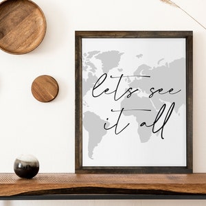 May include: A framed print featuring a grey world map and the text "let's see it all" in black script. The print is hanging on a white wall above a wooden table.