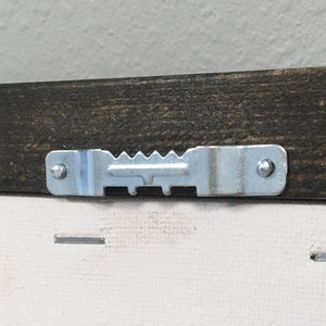 May include: Close-up of the back of a framed canvas painting. The frame is dark brown wood with a silver metal hanging hook. The canvas is white and has a textured surface.