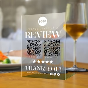 May include: A clear acrylic sign with the text "Please leave us a review" and two QR codes for Google and Facebook. The sign also includes the text "Thank you!" and social media handles.