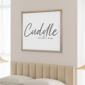 May include: A bedroom scene featuring a bed with a beige headboard, white pillows, and a beige blanket. A framed artwork with the text "Cuddle with me" hangs above the bed. The room includes a patterned rug and decorative hanging lights.