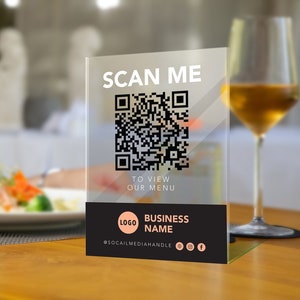 May include: A black and white QR code on a clear acrylic sign with the text "SCAN ME" to view our menu. The sign also includes the text "BUSINESS NAME" and social media icons.