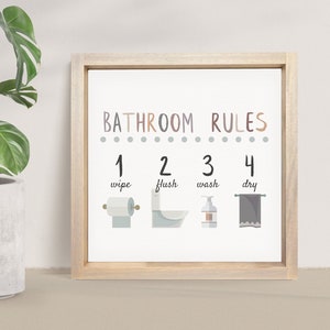May include: A light wood framed print with the text "Bathroom Rules" in brown and gray lettering. The print has four steps for bathroom etiquette, illustrated with icons: 1. wipe, 2. flush, 3. wash, 4. dry.