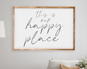May include: A framed print with the text "This is our happy place" in a white frame with a wood-toned border. The print is hanging on a white wall above a white couch with a green throw blanket and a book on top.