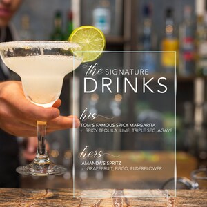 May include: A clear glass margarita with a salt rim and a lime wheel garnish, positioned next to a transparent acrylic sign that reads: the SIGNATURE DRINKS, his TOM'S FAMOUS SPICY MARGARITA - SPICY TEQUILA, LIME, TRIPLE SEC, AGAVE, hers AMANDA'S SPRITZ - GRAPEFRUIT, PISCO, ELDERFLOWER.