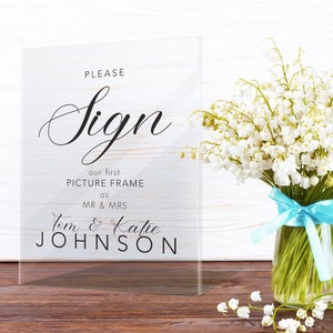 May include: A clear acrylic sign with the text "Please Sign our first picture frame as Mr & Mrs Tom & Katie Johnson". The sign is sitting on a wooden surface with a bouquet of white flowers in a glass vase.