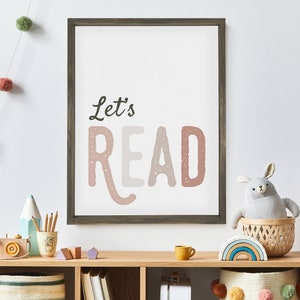 May include: A framed print with the text "Let's Read" in a pink, white, and gray color scheme. The print is hanging on a white wall above a wooden shelf with various children's toys and books.