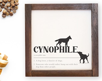 May include: A framed print with a brown wood frame and a white background. The print features the word "CYNOPHILE" in black text with a definition below. The definition reads: "(cy-no-phile) noun 1. A dog lover, a fancier of dogs. 2. Someone who would rather hang out with their dog than other people."