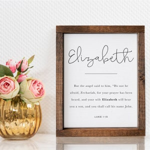 May include: A framed print with a white background and black text. The text reads "Elizabeth" in cursive at the top and a Bible verse from Luke 1:13 below. The frame is made of dark wood.