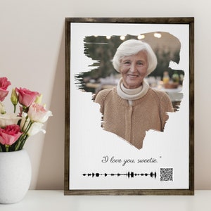 May include: A framed print with a black and white photo of an older woman smiling. The photo is surrounded by a white brushstroke effect. The text "I love you, sweetie." is below the photo, along with a black and white audio waveform and a QR code.