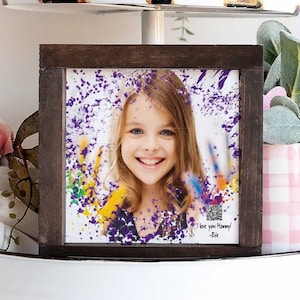 May include: A framed photo of a smiling girl with colorful paint splatters. The dark brown wooden frame sits on a white tiered tray with decorative items, including a pink flower, rolling pins, and a pink and white gingham container.