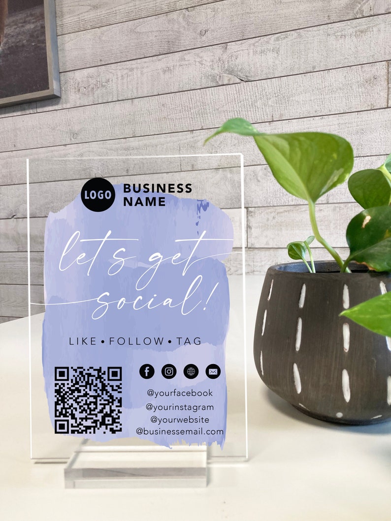 May include: A clear acrylic sign with a purple watercolour background and the text "let's get social!"  The sign also includes a QR code, social media icons, and the text "LIKE FOLLOW TAG" along with social media handles and an email address.