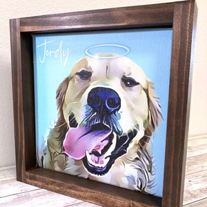 May include: A framed canvas print of a golden retriever dog with a halo above its head. The dog is looking at the viewer with its tongue sticking out. The canvas is on a light blue background and the frame is brown wood. The name "Jordy" is written in white cursive above the dog's head.
