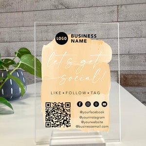 May include: A clear acrylic sign with a watercolour-style background in shades of peach and orange. The text "let's get social!" is written in black script font. The sign also includes a QR code, social media icons, and the text "LIKE FOLLOW TAG" along with social media handles and an email address.