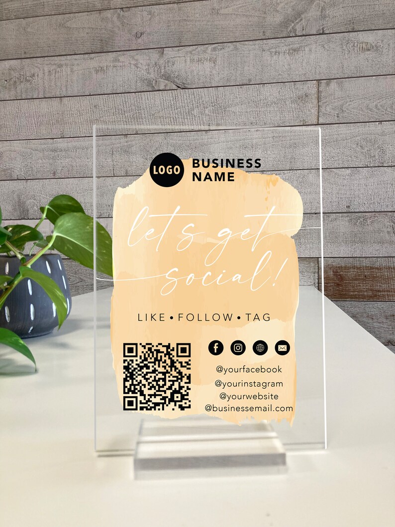 May include: A clear acrylic sign with a watercolour-style background in shades of peach and orange. The text "let's get social!" is written in black script font. The sign also includes a QR code, social media icons, and the text "LIKE FOLLOW TAG" along with social media handles and an email address.