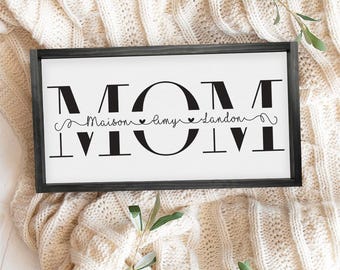 May include: A white sign with black lettering that reads "MOM" with the names "Maison", "Amy", and "Landon" written underneath in a cursive font. The sign is framed in a dark gray frame.