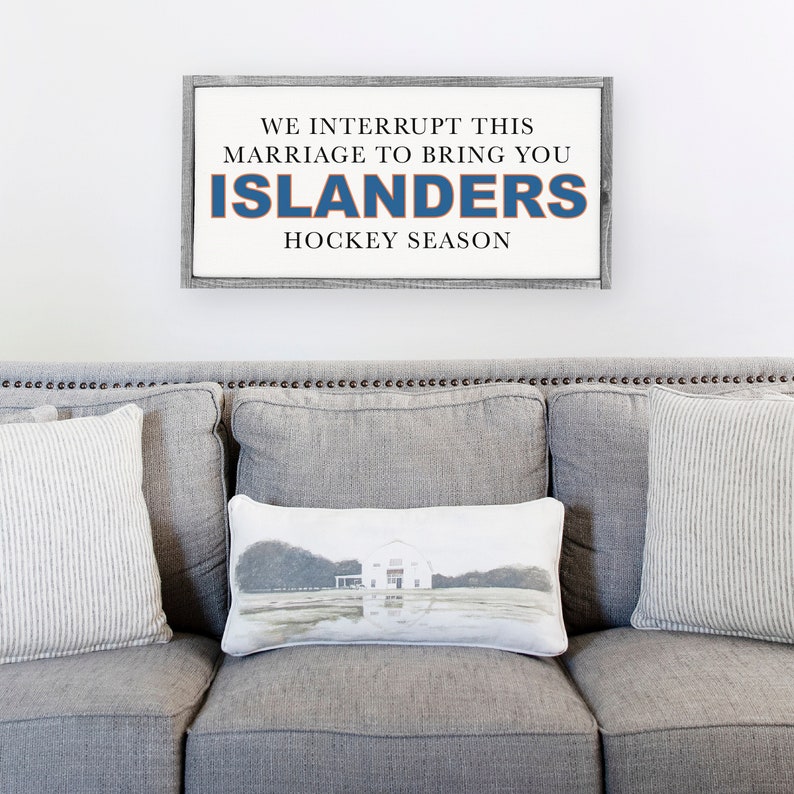 Pu&ograve; includere: Un cartello di legno grigio con testo bianco che recita "We interrupt this marriage to bring you Islanders Hockey Season". Il testo "Islanders" &egrave; in blu. Il cartello &egrave; appeso a una parete bianca sopra un divano grigio con cuscini.