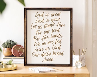 Puede incluir: Un letrero de madera enmarcado con fondo blanco y texto marrón. El texto dice "Dios es grande Dios es bueno Démosle gracias Por nuestra comida Por sus manos Todos somos alimentados Danos Señor Nuestro pan de cada día Amén".