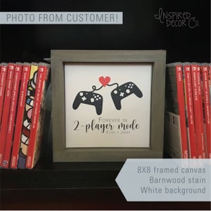 May include: A framed canvas print with a white background and a black and red design of two video game controllers connected by a red heart. The text "Forever in 2-player mode Elise + Jamie" is printed below the controllers. The frame is 8x8 inches and is made of barnwood.
