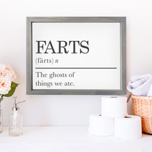 May include: A framed print with a humorous definition of the word "farts" in a black and white font. The definition reads "(fārts) n. The ghosts of things we ate."