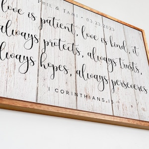 May include: A white wooden sign with a brown wood frame. The sign has black calligraphy text that reads "love is patient, love is kind, it always protects, always trusts, always hopes, always perseveres I Corinthians.13".