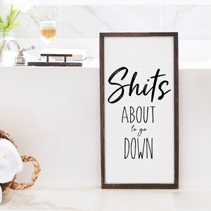 A 12x24 framed canvas stands upright against a bathtub. The text on the canvas reads, "Shit's about to go down".