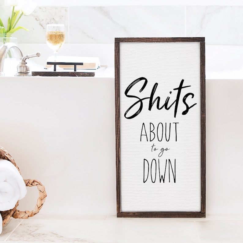 A 12x24 framed canvas stands upright against a bathtub. The text on the canvas reads, "Shit's about to go down".