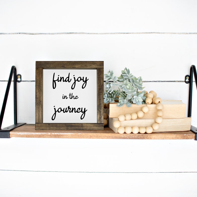 May include: A wooden framed sign with the text "find joy in the journey" in black script. The sign is on a wooden shelf with books, faux succulents, and a wooden bead garland. The shelf is supported by black metal brackets.