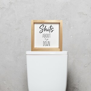 Pode incluir: Uma placa enquadrada em madeira com o texto "Shits About to go Down" em um fundo branco. A placa est&aacute; em cima de um vaso sanit&aacute;rio branco.