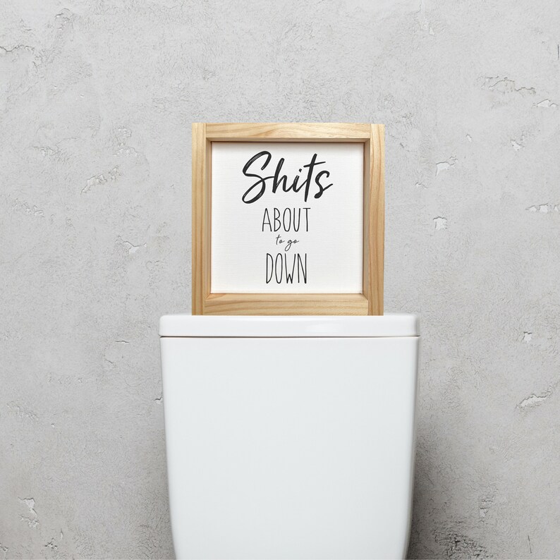 Pode incluir: Uma placa enquadrada em madeira com o texto "Shits About to go Down" em um fundo branco. A placa est&aacute; em cima de um vaso sanit&aacute;rio branco.