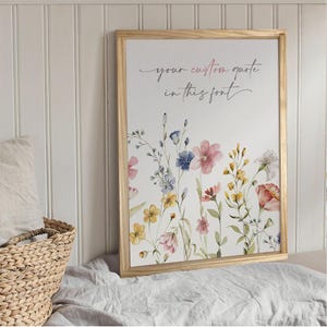 May include: A framed print with a watercolour floral design featuring pink, blue, and yellow flowers. The text "your custom quote in this font" is written in a cursive font at the top of the print.