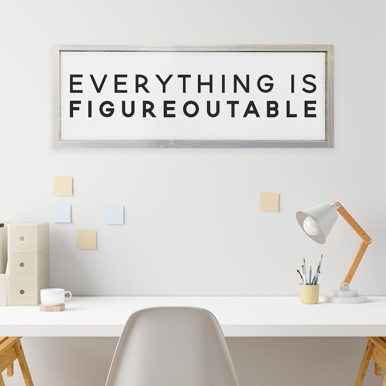 May include: A white framed sign with the text "EVERYTHING IS FIGUREOUTABLE" in black lettering. The sign is hanging on a white wall above a white desk with a lamp, a cup, and a chair.
