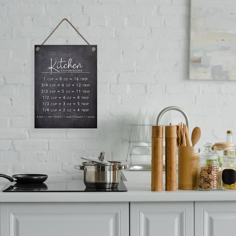 May include: Black chalkboard sign with white text that reads "Kitchen conversions" and lists various measurements for cups, ounces, and tablespoons. The sign is hanging from a string and is displayed in a kitchen setting.