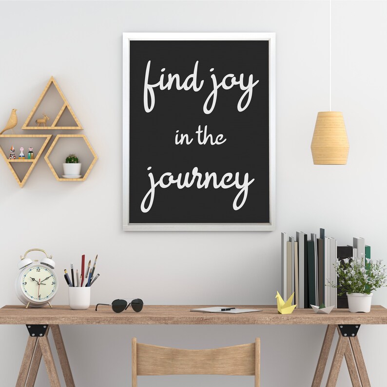 May include: A framed print with the words "find joy in the journey" in white script on a black background. The print hangs above a wooden desk with a clock, sunglasses, and books. A wooden shelf with geometric shapes is on the wall.