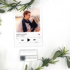 May include: A white card with a photo of a couple in a winter setting. The card is designed to look like a music player with the song title "Speechless" by Dan + Shay. A red heart icon is in the top right corner. A small white card with a QR code and the text "Our Forever Song" is below the larger card.