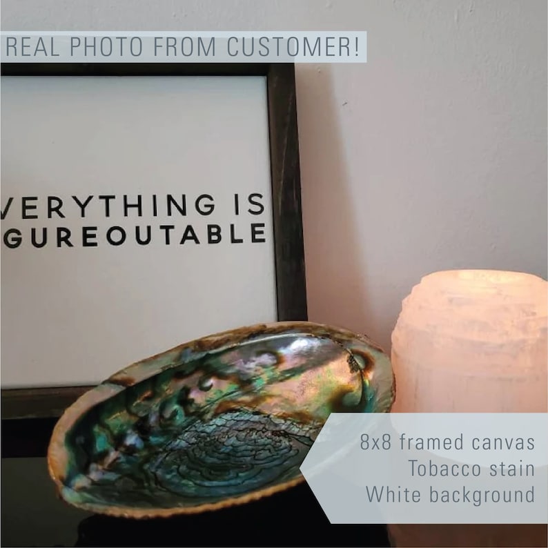 May include: An 8x8 inch framed canvas with the text "EVERYTHING IS FIGUREOUTABLE" in black lettering on a white background. The canvas is framed in a dark wood frame. The canvas is sitting on a table with a seashell and a white candle.