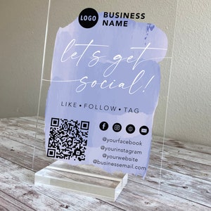 May include: A clear acrylic sign with a purple watercolour background. The text "let's get social!" is written in white. The sign also includes a QR code, social media icons, and the text "LIKE FOLLOW TAG" along with social media handles and an email address.