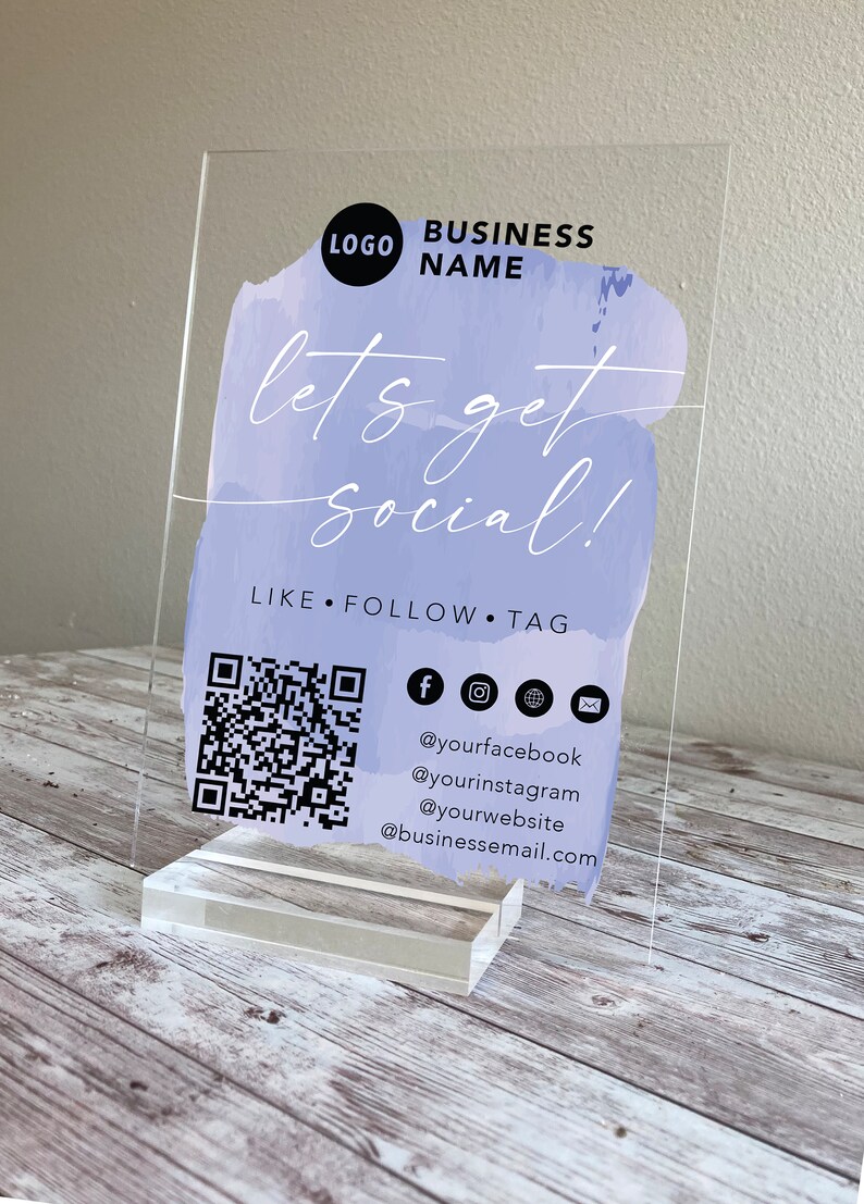 May include: A clear acrylic sign with a purple watercolour background. The text "let's get social!" is written in white. The sign also includes a QR code, social media icons, and the text "LIKE FOLLOW TAG" along with social media handles and an email address.