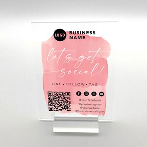 May include: A white acrylic sign with a pink watercolour background. The sign says "let's get social!" and includes a QR code, social media icons, and the text "LIKE FOLLOW TAG".