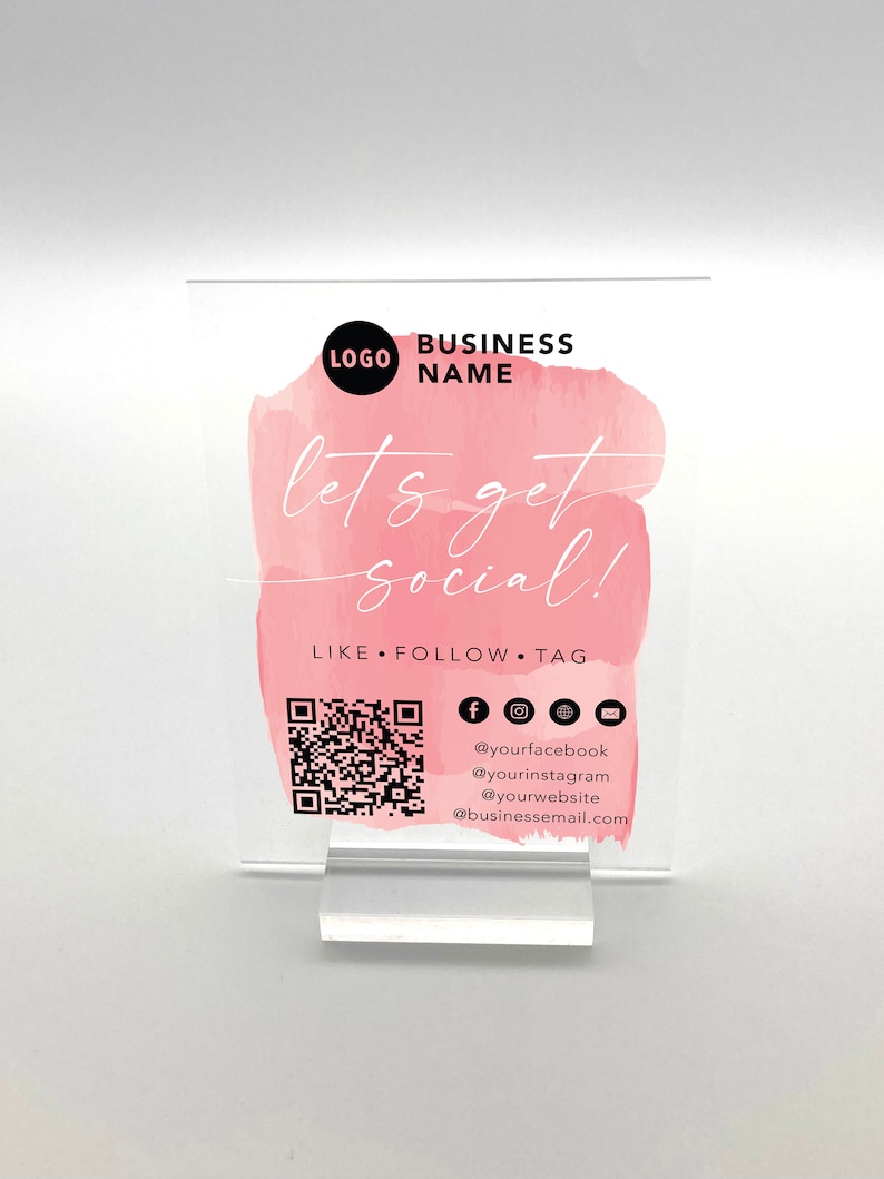 May include: A white acrylic sign with a pink watercolour background. The sign says "let's get social!" and includes a QR code, social media icons, and the text "LIKE FOLLOW TAG".