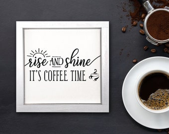 May include: A white framed sign with the text "rise and shine it's coffee time" in black lettering. The sign is on a dark gray background with a cup of coffee and coffee beans.