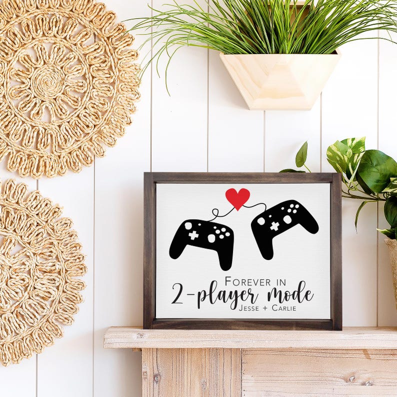 May include: A white sign with a black border and a black and white illustration of two video game controllers connected by a red heart. The sign reads "Forever in 2-player mode Jesse + Carlie".