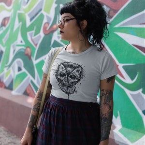 May include: A woman wearing a white t-shirt with a black and white graphic print of a skull with two faces. She is wearing a black and red plaid skirt and has a black leather jacket slung over her shoulder.