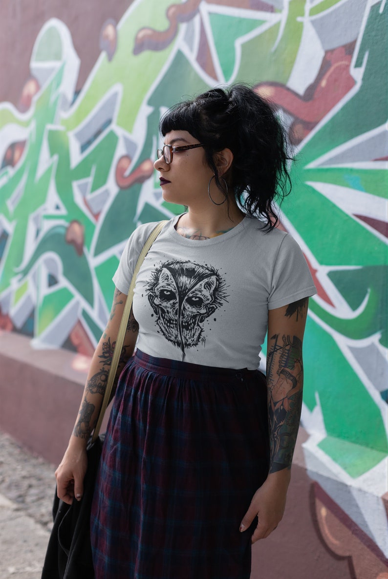May include: A woman wearing a white t-shirt with a black and white graphic print of a skull with two faces. She is wearing a black and red plaid skirt and has a black leather jacket slung over her shoulder.