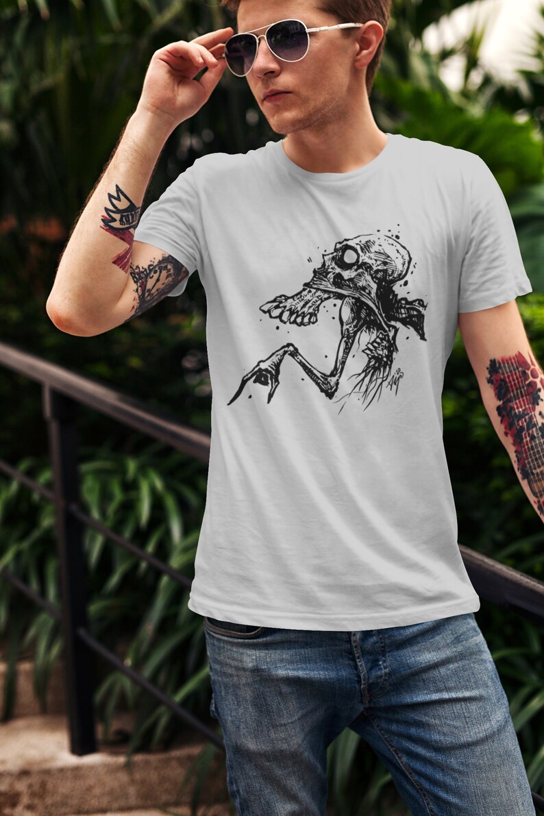 Peut inclure: T-shirt gris clair avec une illustration en noir et blanc d'une figure squelettique. Le motif graphique est centr&eacute; sur le t-shirt et repr&eacute;sente une cr&eacute;ature avec un cr&acirc;ne et des membres osseux. Le t-shirt est en mati&egrave;re souple.