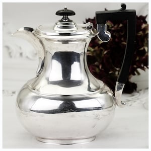 May include: A silver coffee pot with a black handle and lid knob. The pot has a rounded body, a curved spout, and a lid with a small, round knob. The handle is a dark, curved shape. The pot is reflective and appears to be made of metal.