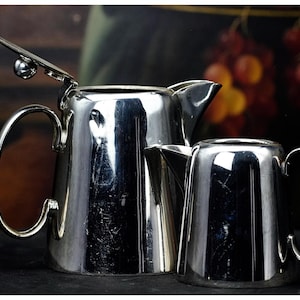 May include: Two polished silver teapots, one larger with a hinged lid and a curved handle, and a smaller one with a similar design. The reflective surfaces create a bright, elegant appearance. The background is dark with blurred fruit.