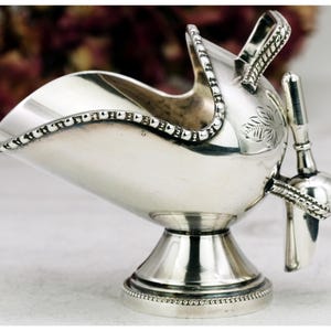 May include: A silver sugar scuttle with a small scoop. The scuttle has a decorative rim and a handle. The scoop is attached to the side of the scuttle. The scuttle sits on a pedestal base. The design includes a leaf pattern.