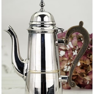 Puede incluir: Una cafetera plateada pulida con un asa decorativa. La cafetera tiene un pico largo y una tapa con un pequeño adorno. El asa es de color marrón oscuro. La cafetera es probablemente de acero inoxidable.