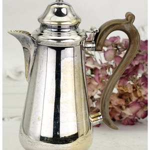 May include: A silver coffee pot with a decorative wooden handle. The pot features a rounded base, a long spout, and a detailed lid. The handle curves elegantly, enhancing the vintage appeal. A classic item for serving beverages.