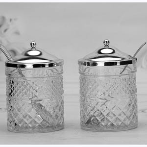 May include: Two clear glass jars with silver lids and small spoons. The jars have a diamond pattern and are designed for serving condiments. The lids have a small knob on top.